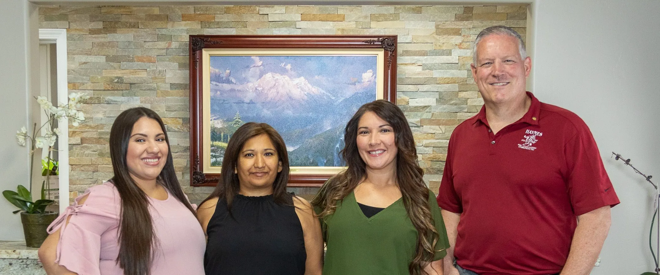 Four staff members standing together in front of a stone wall with a framed mountain landscape photo at Haynes Chiropractic