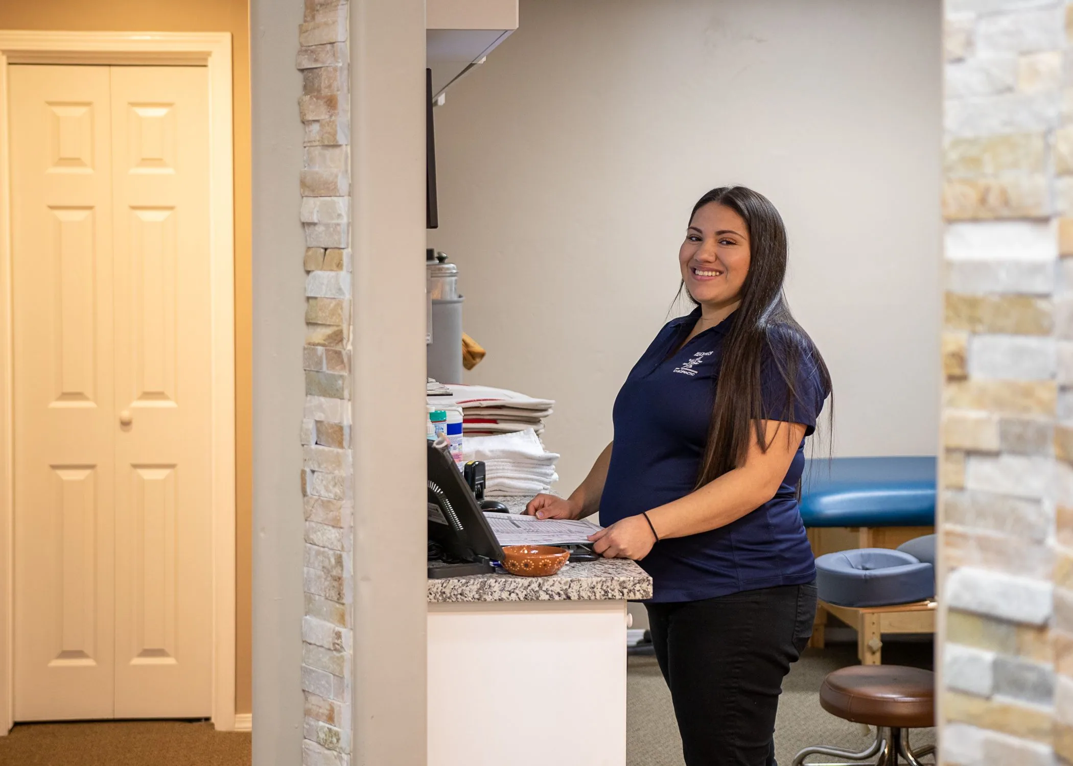 Woman in navy polo shirt standing at reception desk in medical office with brick columns and yellow door