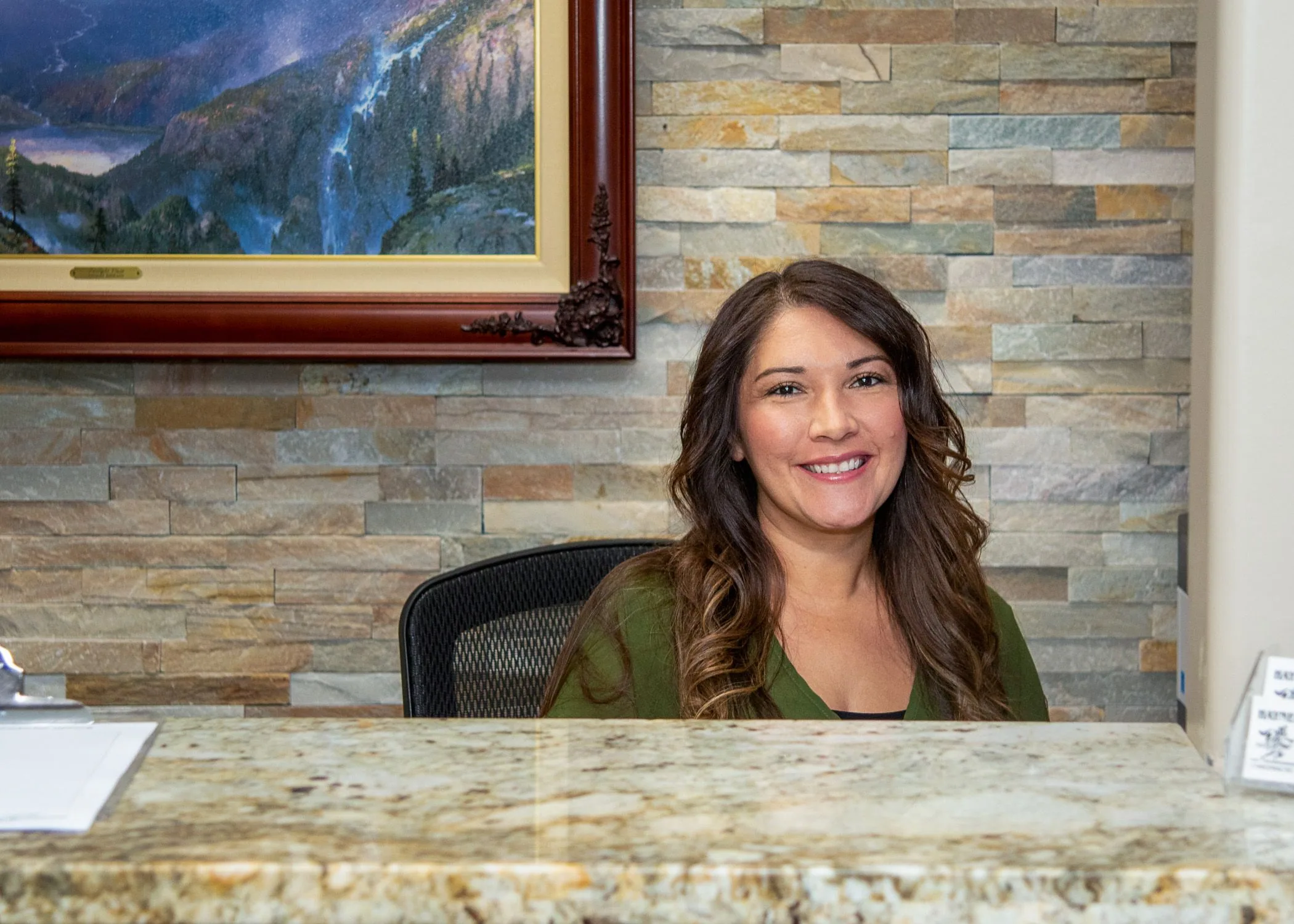 Woman with long brown hair wearing olive green jacket seated at granite desk in office with stone wall and framed mountain landscape