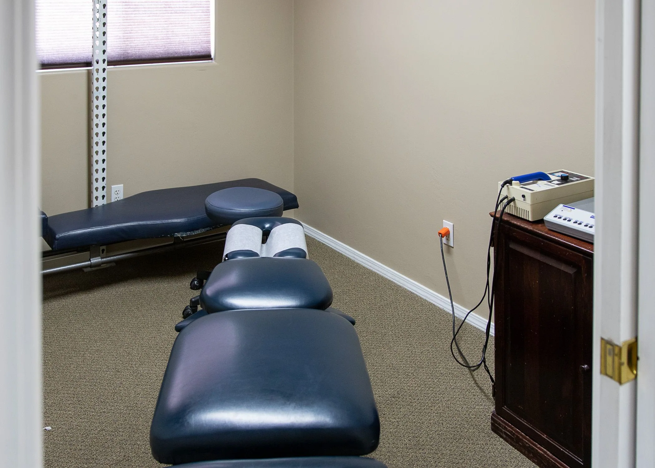 Chiropractic treatment room with blue padded adjustment tables, equipment on shelves, and a window with blinds