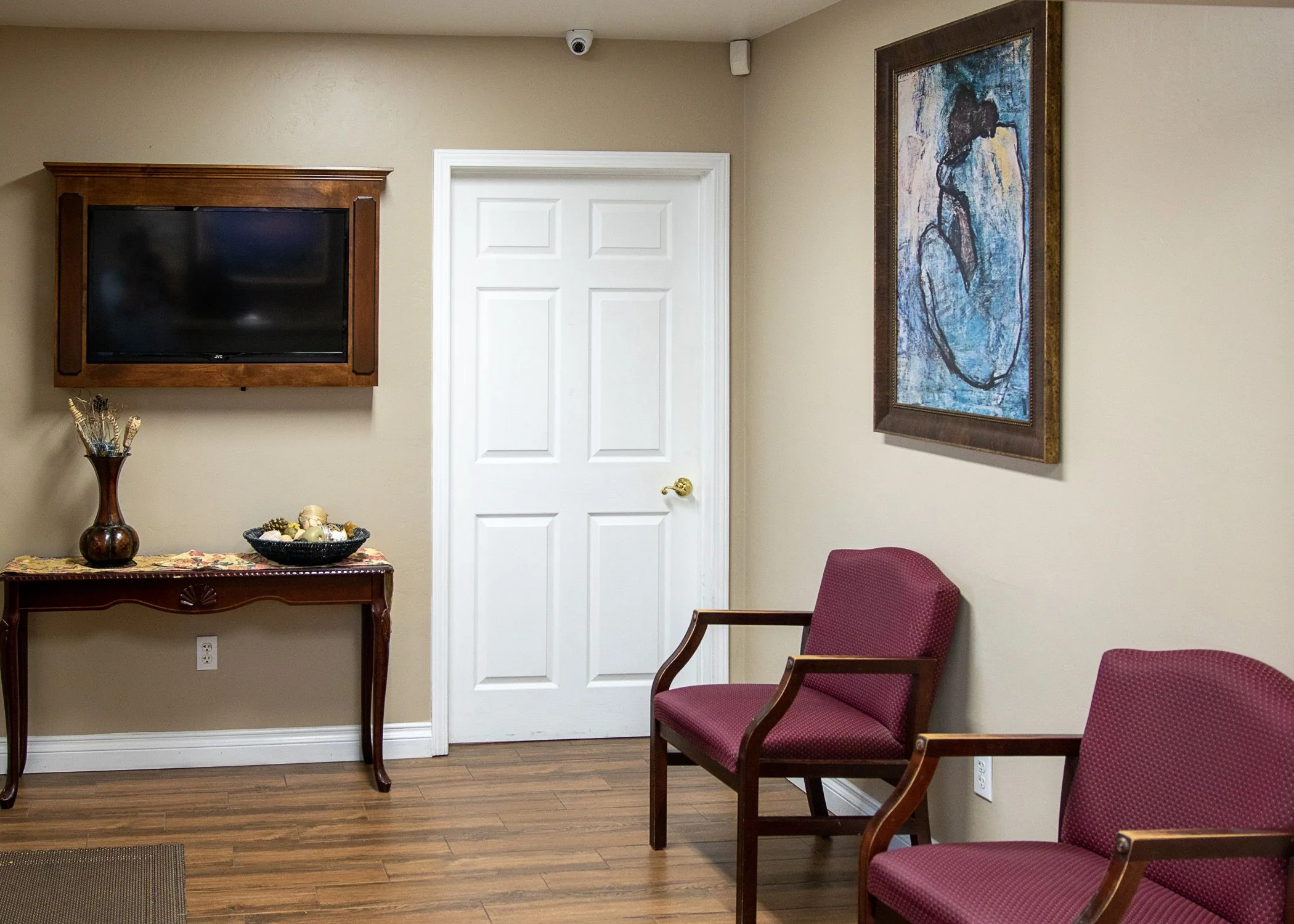 Medical waiting room with burgundy chairs, mounted television, white door, decorative wall art, and wooden furniture